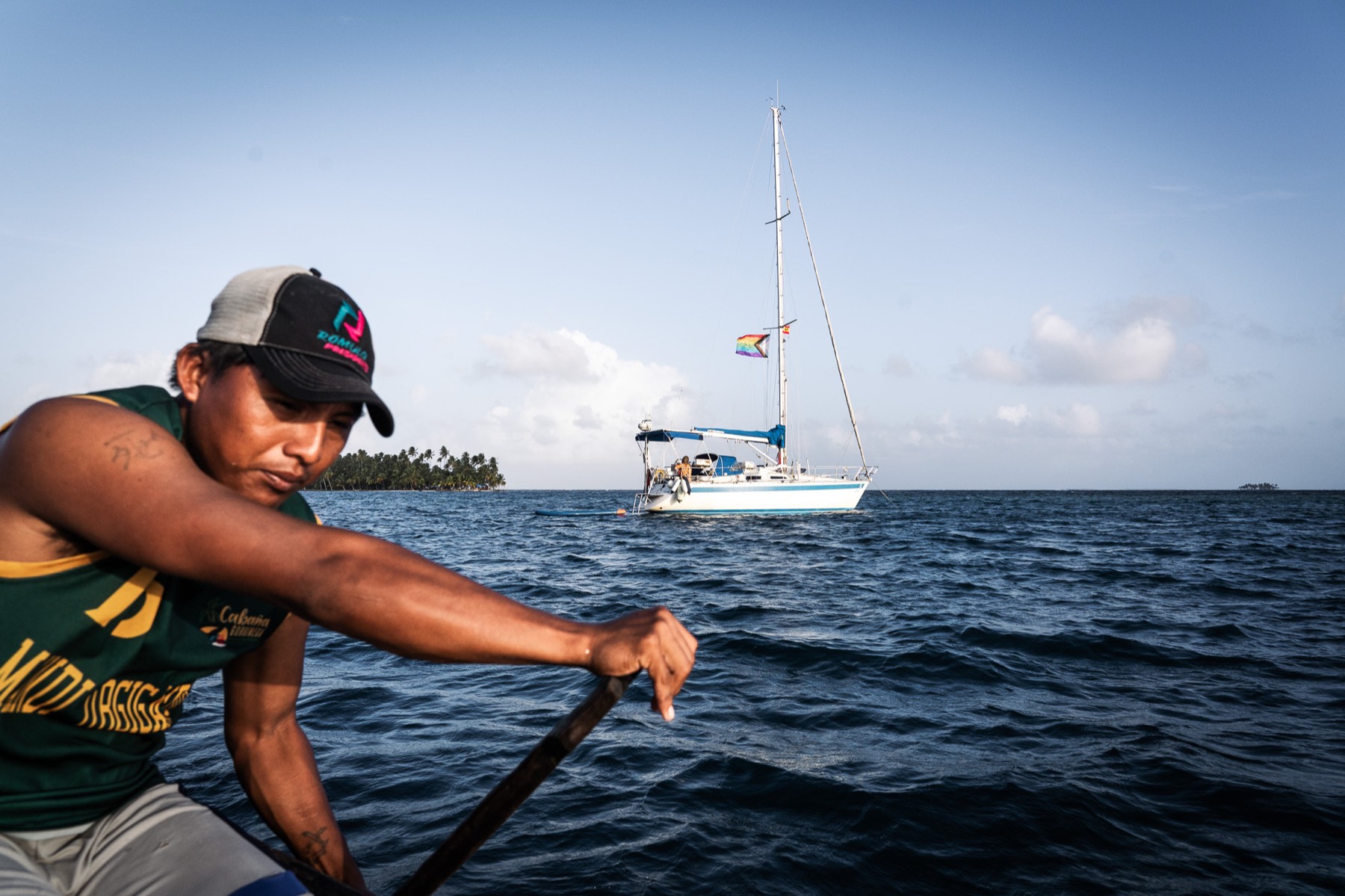 A Guna man paddles a dugout canoe with the sailboat Falkor anchored behind him